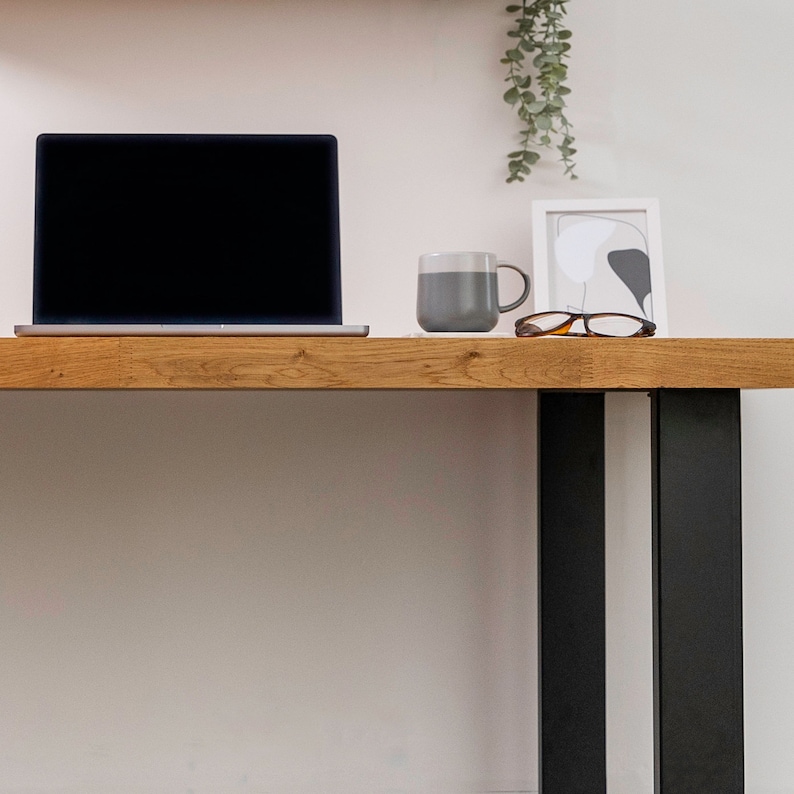 Pu&ograve; includere: Una scrivania in legno con gambe in metallo nero. Un computer portatile, una tazza grigia e un paio di occhiali sono sulla scrivania.