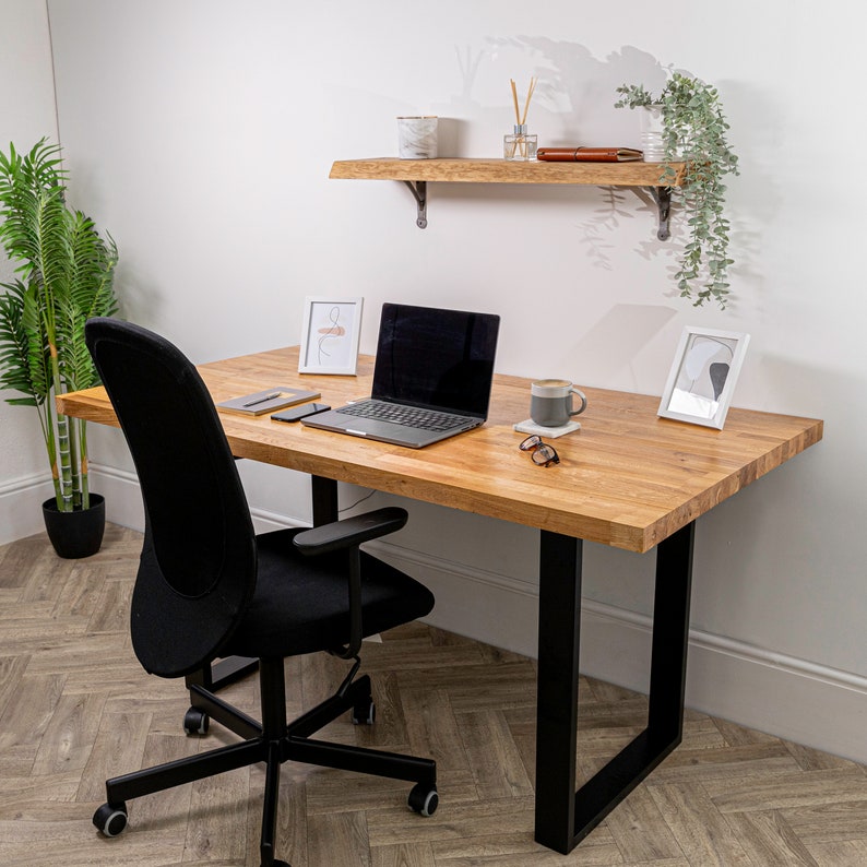 Pu&ograve; includere: Una scrivania in legno chiaro con gambe in metallo nero e una sedia da ufficio nera. Un laptop, una tazza di caff&egrave; e un paio di occhiali da sole sono sulla scrivania. C'&egrave; uno scaffale sopra la scrivania con una piccola pianta e un libro.