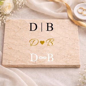 Könnte beinhalten: Eine rechteckige Marmorplatte mit drei Initialen-Sets: "D|B", "D♥B" und "D∞B". Die Platte ist umgeben von weißen Blumen, einem goldenen Band und einer kleinen Schale mit zwei goldenen Ringen. Der Hintergrund ist ein weicher, neutraler Stoff.