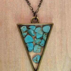 May include: A triangular pendant necklace featuring a bronze-toned metal frame filled with multiple raw, light blue turquoise stones. The pendant hangs from a matching bronze-toned metal chain, showcasing a geometric design with natural stone textures.