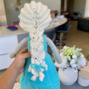 Pode incluir: Boneca de croch&ecirc; feita &agrave; m&atilde;o com um vestido azul claro, cabelo branco e uma longa tran&ccedil;a. O cabelo da boneca &eacute; adornado com enfeites brilhantes. A boneca tem bra&ccedil;os cinzentos e um la&ccedil;o azul claro. &Eacute; um artesanato feito &agrave; m&atilde;o.