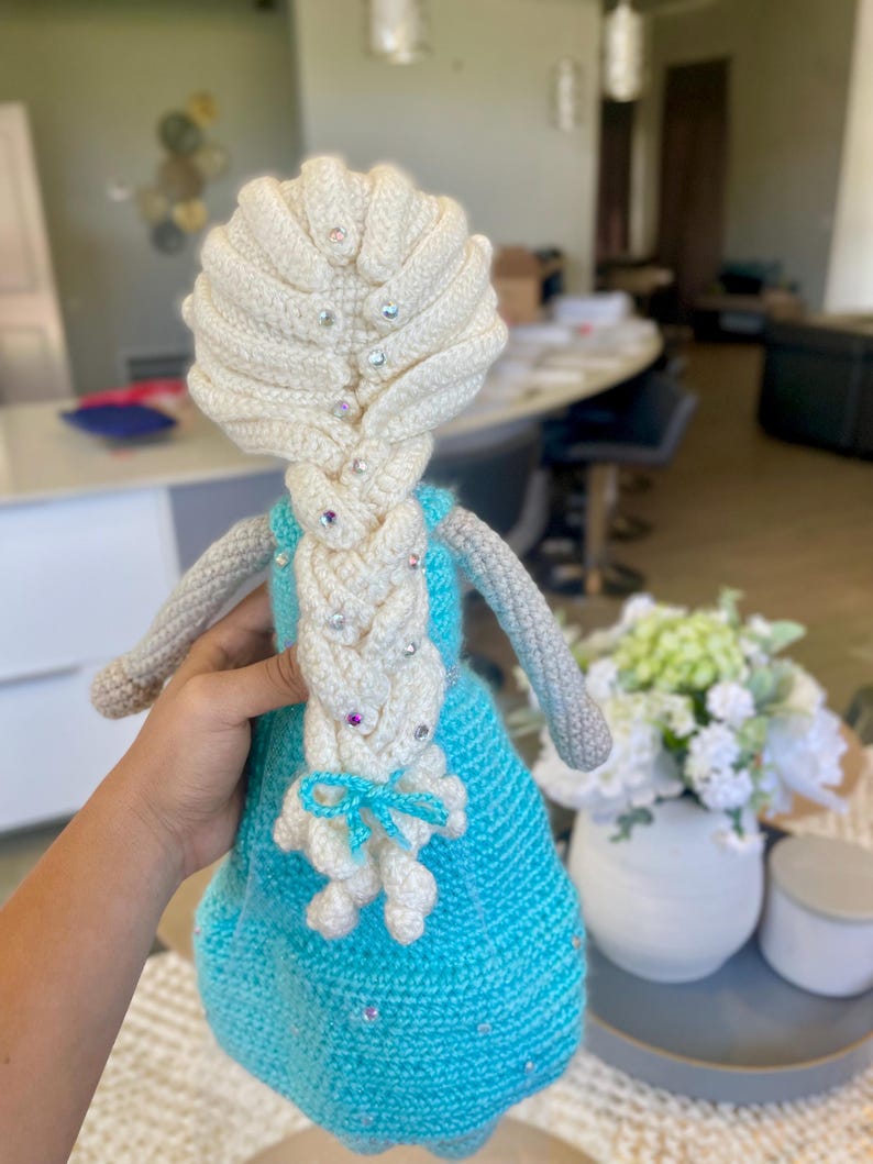 Pode incluir: Boneca de croch&ecirc; feita &agrave; m&atilde;o com um vestido azul claro, cabelo branco e uma longa tran&ccedil;a. O cabelo da boneca &eacute; adornado com enfeites brilhantes. A boneca tem bra&ccedil;os cinzentos e um la&ccedil;o azul claro. &Eacute; um artesanato feito &agrave; m&atilde;o.