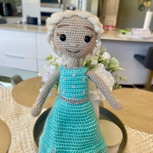 Pode incluir: Boneca de croch&ecirc; feita &agrave; m&atilde;o com tom de pele claro, cabelo loiro e um vestido turquesa. A boneca tem um cinto prateado e detalhes decorativos. A boneca est&aacute; em cima de uma mesa.