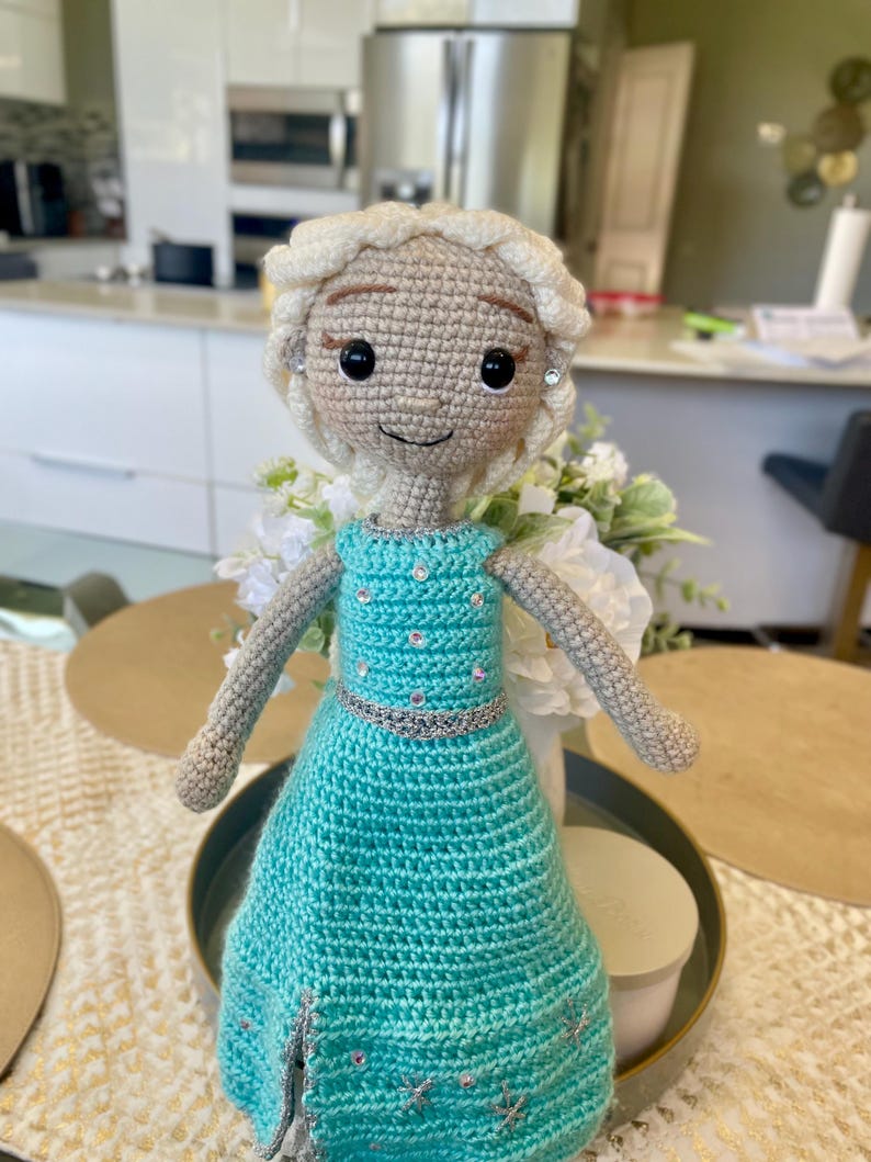 Pode incluir: Boneca de croch&ecirc; feita &agrave; m&atilde;o com tom de pele claro, cabelo loiro e um vestido turquesa. A boneca tem um cinto prateado e detalhes decorativos. A boneca est&aacute; em cima de uma mesa.
