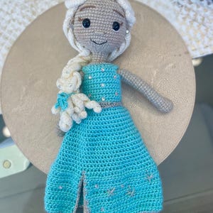 Pode incluir: Boneca de croch&ecirc; com um vestido azul claro com detalhes prateados, um penteado tran&ccedil;ado branco e um pequeno urso branco. A boneca tem olhos pretos e uma express&atilde;o sorridente. O vestido apresenta uma fenda e decora&ccedil;&otilde;es em forma de floco de neve.
