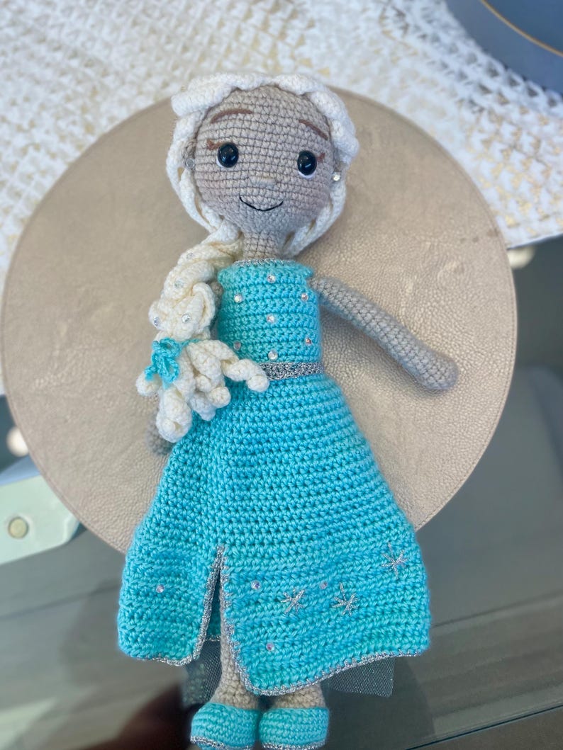 Pode incluir: Boneca de croch&ecirc; com um vestido azul claro com detalhes prateados, um penteado tran&ccedil;ado branco e um pequeno urso branco. A boneca tem olhos pretos e uma express&atilde;o sorridente. O vestido apresenta uma fenda e decora&ccedil;&otilde;es em forma de floco de neve.