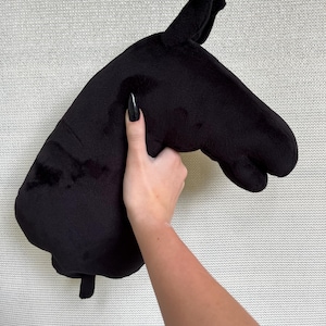 Pode incluir: Uma almofada preta de pelúcia com a forma de uma cabeça de cavalo. A almofada tem a forma de uma cabeça de cavalo com orelhas e focinho. A almofada é feita de um material macio e aveludado.