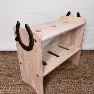 May include: A light-coloured wooden shelf with two tiers and a horseshoe design. The shelf has a rectangular shape with rounded edges and is held together with brass-coloured screws. Two dark metal horseshoes are attached to the sides.