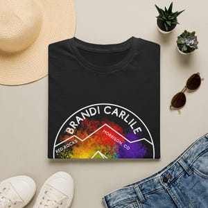 May include: A black t-shirt featuring a white circular outline containing the text Brandi Carlile, Red Rocks, and Morrison, CO. Inside the circle is a colorful, abstract mountain graphic with vibrant rainbow hues of red, orange, yellow, green, and purple.