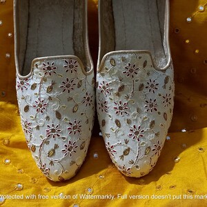 May include: A pair of white embroidered flat shoes with gold and red floral designs. The shoes are on a yellow fabric background.