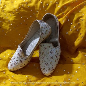 May include: A pair of white embroidered flat shoes with a floral pattern. The shoes have a pointed toe and a closed back. The shoes are on a yellow fabric background.