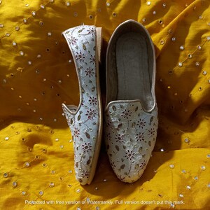 May include: A pair of white embroidered flat shoes with gold and red accents. The shoes are on a yellow fabric background.