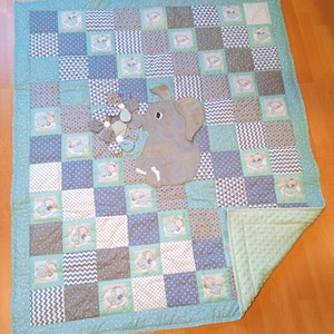 Könnte beinhalten: Eine Babydecke mit Patchwork-Muster in Blau- und Grüntönen. Die Decke hat eine graue Elefanten-Applikation mit Blumenmuster. Die Decke hat eine weiche, plüschige Rückseite.