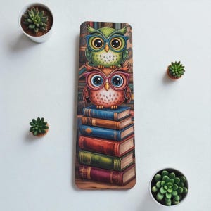 Peut inclure: Marque-page rectangulaire avec deux hiboux de dessin animé portant des lunettes. Les hiboux sont assis sur une pile de livres. Le hibou du haut est vert avec des lunettes bleues, et le hibou du bas est orange avec des lunettes roses. Les livres sont de couleurs variées.