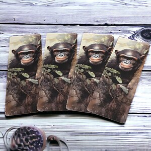 May include: Four rectangular bookmarks featuring a chimpanzee portrait. The chimpanzees have dark fur and are set against a backdrop of tree bark and foliage. The bookmarks are arranged on a light wood-grain surface.
