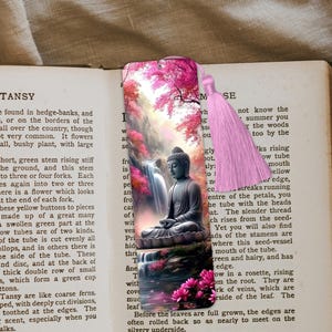 May include: A pink and green bookmark with a tassel. The bookmark features a painting of a meditating Buddha in a forest setting with a waterfall. The bookmark has the text "TANSY" printed on it.