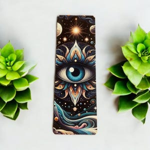 May include: A bookmark with a colorful, psychedelic design featuring a large eye surrounded by a mandala-like pattern. The background is a dark blue with stars and planets.