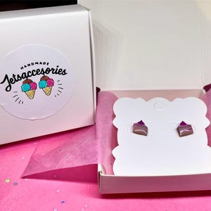 May include: A pair of cake slice stud earrings displayed in a white gift box with pink tissue paper. The earrings are brown, purple, and white. The box has a sticker with the text "Handmade Jetsaccessories" and ice cream cones.