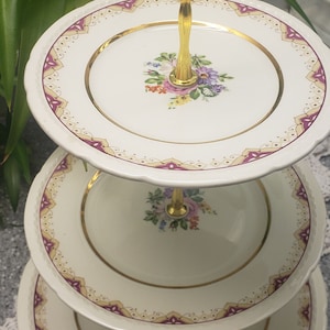 May include: A three-tiered serving stand with a gold handle and trim. Each plate is white with a floral design and a burgundy and gold border. The stand is ideal for displaying desserts or appetizers.