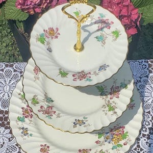Könnte beinhalten: Ein dreistöckiger Kuchenständer mit goldenem Griff. Die weißen Teller haben ein Blumenmuster in Rosa, Blau und Gelb mit goldenem Rand. Der Ständer steht auf einer weißen Spitzendecke.