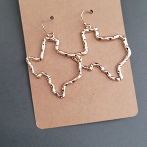 May include: A pair of gold-tone earrings shaped like the state of Texas. The earrings are textured and have a hammered finish.