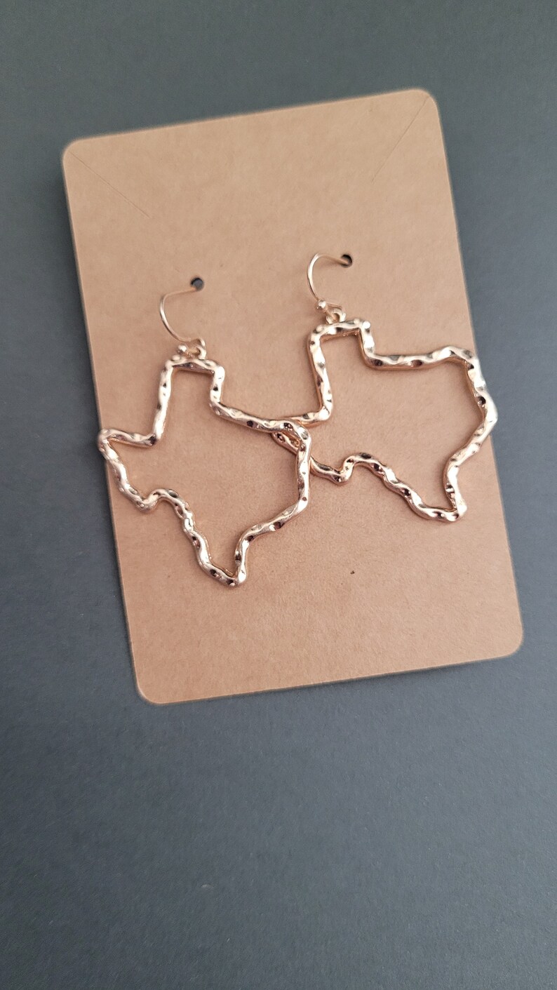 May include: A pair of gold-tone earrings shaped like the state of Texas. The earrings are textured and have a hammered finish.