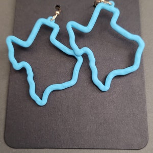 May include: A pair of blue Texas-shaped earrings. The earrings are made of a smooth, pliable material and have a gold-colored hook.