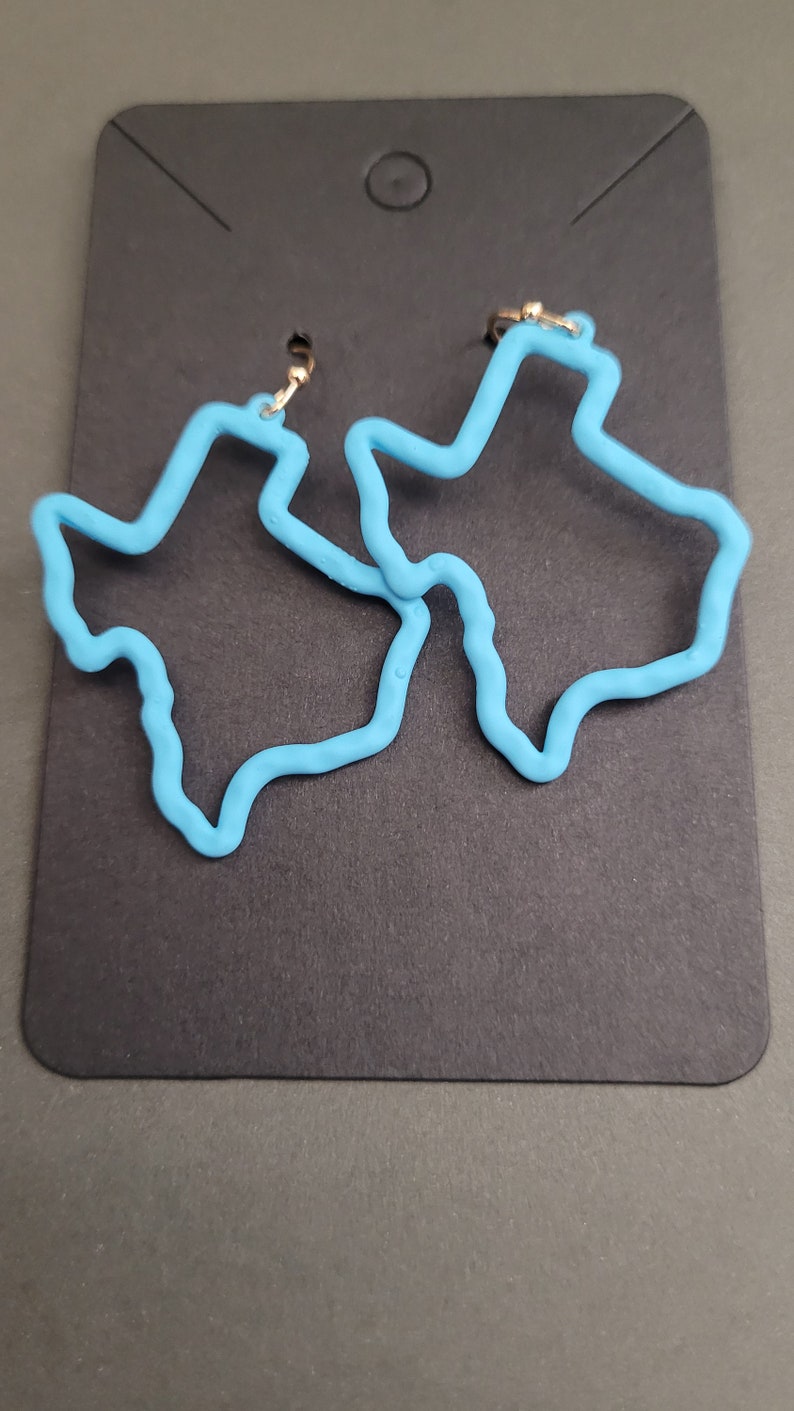 May include: A pair of blue Texas-shaped earrings. The earrings are made of a smooth, pliable material and have a gold-colored hook.