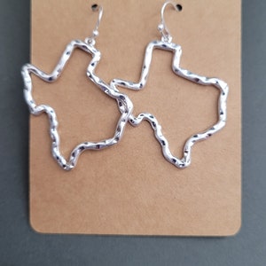 May include: A pair of silver earrings shaped like the outline of the state of Texas. The earrings are made of twisted wire and have a textured finish.