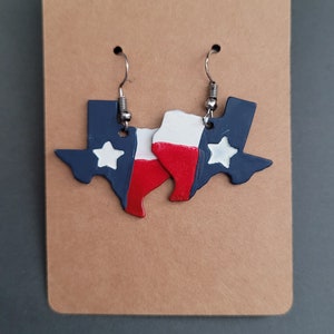 May include: A pair of earrings shaped like the state of Texas, painted in the colors of the Texas flag: blue, white, and red. The earrings have silver hooks.