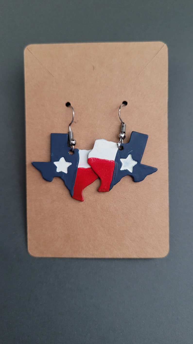 May include: A pair of earrings shaped like the state of Texas, painted in the colors of the Texas flag: blue, white, and red. The earrings have silver hooks.