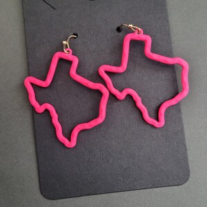 May include: A pair of pink Texas-shaped earrings with gold hooks. The earrings are on a black background.