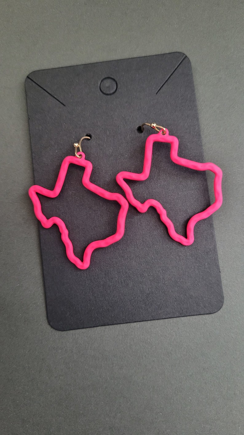 May include: A pair of pink Texas-shaped earrings with gold hooks. The earrings are on a black background.