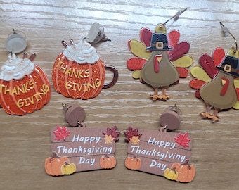 May include: A set of six Thanksgiving-themed earrings. Two pairs feature pumpkin-shaped earrings with the words "Thanks Giving" on them. Another pair features a brown turkey with a black hat and red and yellow feathers. The final pair features a wooden sign with the words "Happy Thanksgiving Day" on it.