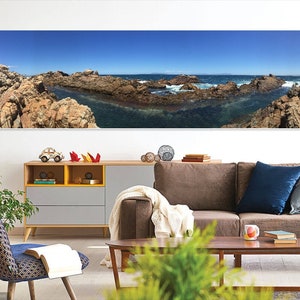 May include: A panoramic photograph of a rocky coastline with blue water and a bright blue sky. The photo is hanging on a white wall above a brown sofa with blue pillows.