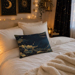 May include: A decorative pillow with a dark blue background featuring a celestial design of clouds, stars, and gold accents. The pillow is rectangular and rests on a bed with white linens and a cream-colored throw blanket.