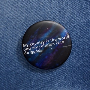 Puede incluir: Un botón negro con un fondo de galaxia y el texto "My country is the world and my religion is to do good."