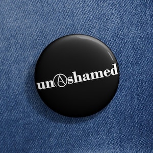Puede incluir: Un botón negro con la palabra "un@shamed" en letras blancas. El botón es redondo y brillante. El botón está sobre una chaqueta vaquera.