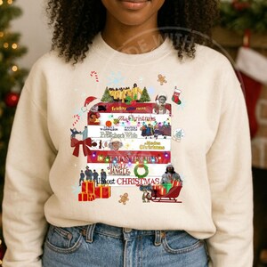 Puede incluir: Sudadera crema con una pila de títulos de películas navideñas, incluyendo "Friday Next" y "This Christmas". Gráficos festivos como un gorro de Papá Noel, hombre de jengibre y regalos envueltos decoran el diseño.