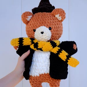 May include: A brown crocheted bear wearing a black witch hat, a white crocheted jacket, and a yellow and black striped scarf.