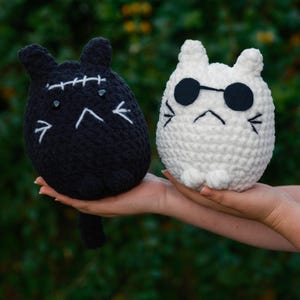 May include: Two crocheted cat plushies, one black with stitched eyes and a stitched mouth, and one white with black sunglasses and a stitched frown.