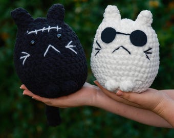 May include: Two crocheted cat plushies, one black with stitched eyes and a stitched mouth, and one white with black sunglasses and a stitched frown.