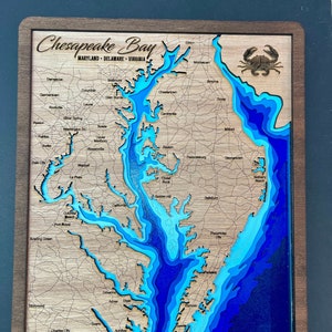 May include: A wooden map of the Chesapeake Bay with a blue, teal, and turquoise layered design. The map includes the states of Maryland, Delaware, and Virginia. The text "Chesapeake Bay" is written in cursive at the top of the map. A small crab is carved into the wood in the upper right corner of the map.