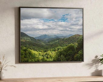 Könnte beinhalten: Ein gerahmter Landschaftsdruck, der eine üppig grüne Bergkette unter einem bewölkten Himmel mit blauen Flecken zeigt. Das Kunstwerk zeigt einen dichten Wald im Vordergrund, mit sanften Hügeln und fernen Gipfeln. Der dunkle Rahmen kontrastiert mit den lebendigen Farben der Szene.