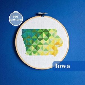 May include: A cross-stitch embroidery hoop featuring the state of Iowa, rendered in a geometric pattern of green, yellow, and blue triangles. The word "Iowa" is displayed in white text on a blue banner. A circular "PDF pattern" graphic is in the upper left.