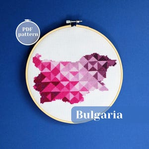 Peut inclure: Un tambour à broder circulaire en bois contenant un tissu Aida blanc avec un motif au point de croix représentant la carte de la Bulgarie. La carte est composée de triangles géométriques dans des tons de rose, magenta et violet foncé. Texte : PDF pattern.
