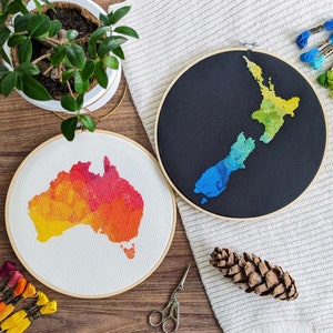 May include: Two embroidery hoops with cross-stitch designs of Australia and New Zealand. The Australian design is a watercolor-style orange and yellow, while the New Zealand design is a blue and green watercolor-style.
