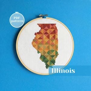 May include: A cross-stitch pattern of the state of Illinois, featuring a geometric design in shades of red, orange, yellow, and green. The design is set within a wooden embroidery hoop. The text "Illinois" is displayed below the hoop.