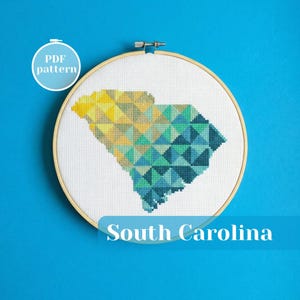 May include: A cross-stitch design of the South Carolina state outline, filled with geometric shapes in yellow, green, and blue. The design is set within a wooden embroidery hoop. Text reads "South Carolina" and "PDF pattern".