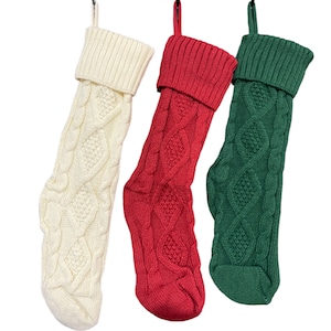 May include: Three knitted Christmas stockings hanging from hooks. The stockings are white, red, and green with a cable knit pattern.